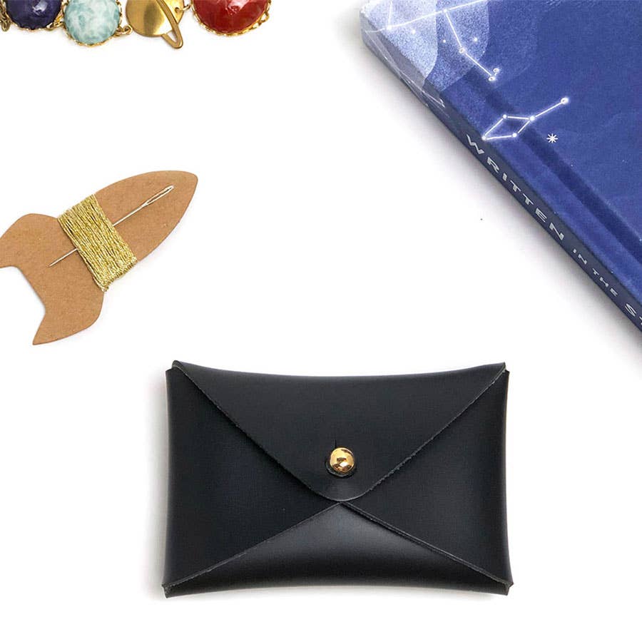 A small black envelope-style pouch or card case with a gold-tone snap button, photographed on a white background with styled props including a blue book, jewelry, and a cardboard rocket.