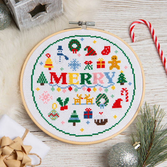 A round wooden embroidery hoop containing a completed cross-stitch design with the word “MERRY” in the center and festive holiday motifs around the border, styled on a light wood surface with candy canes, pine branches, ornaments, and a gift box.