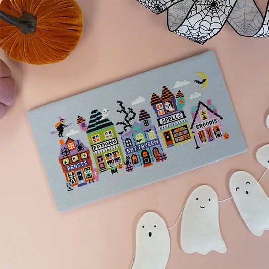 Cross-stitched Halloween-themed fabric panel with colorful buildings on a pink background.