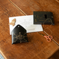 A black envelope-style star sign kit with a gold celestial design, shown on a rustic wooden table alongside a white instruction sheet, a cardboard rocket, peach-handled scissors, and the black product box labeled 'STITCH YOUR STAR SIGN'.