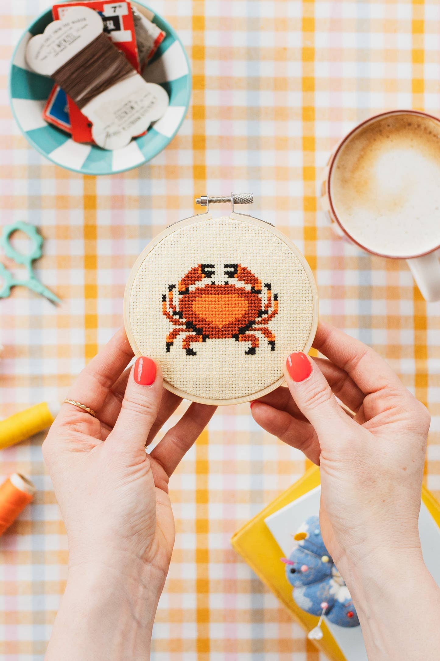 Person holding a cross-stitched crab design on a checkered tablecloth with coffee and craft supplies.