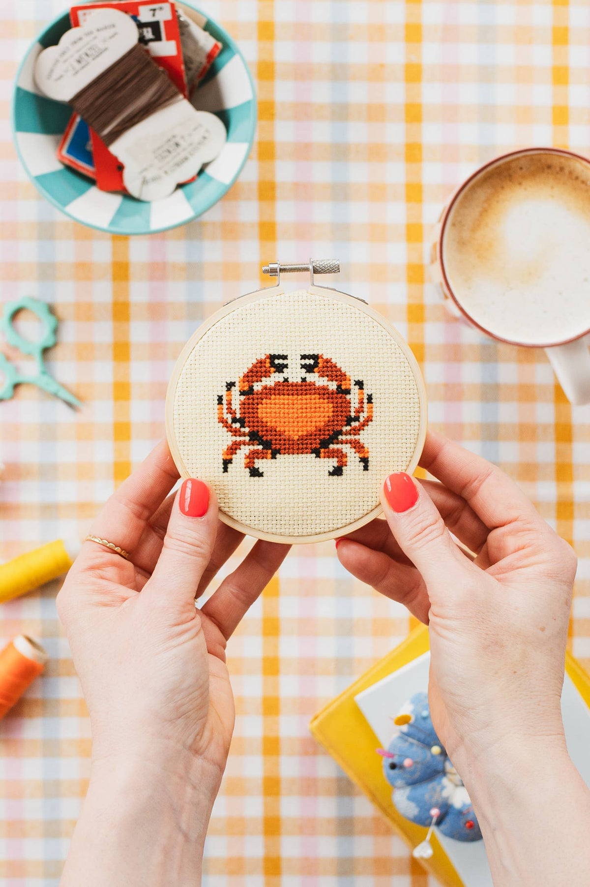 Person holding a cross-stitched crab design on a checkered tablecloth with coffee and craft supplies.