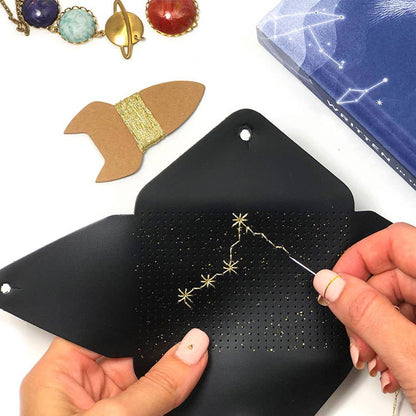 A close-up of hands using a star constellation needlepoint canvas. The black canvas is laid open on a white surface, with a gold needle and thread forming a constellation.