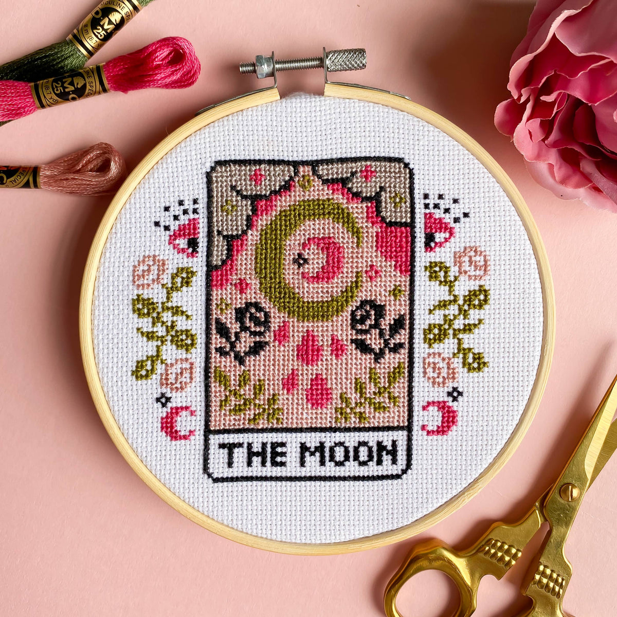 Cross-stitched design of 'The Moon' tarot card with embroidery hoop, threads, and scissors on a pink background.