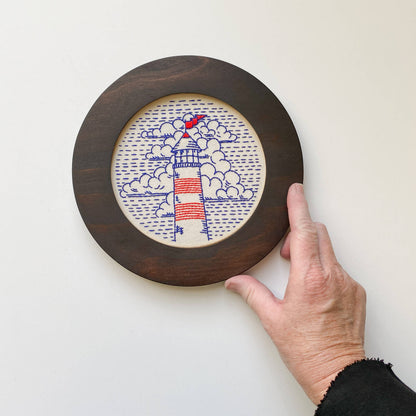 Hand holding a circular wooden embroidery cross stitch frame with a embroidered lighthouse design on a white background