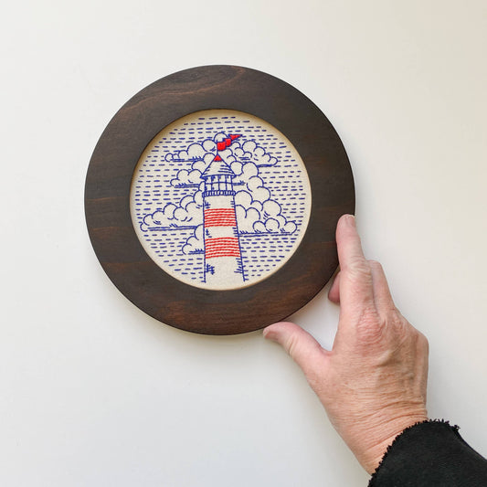 Hand holding a circular wooden embroidery cross stitch frame with a embroidered lighthouse design on a white background