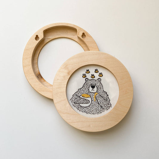 Round light wood cross stitch and embroidery frame with a central white insert featuring a black-and-white bear illustration holding yellow honey, shown on a plain light background.