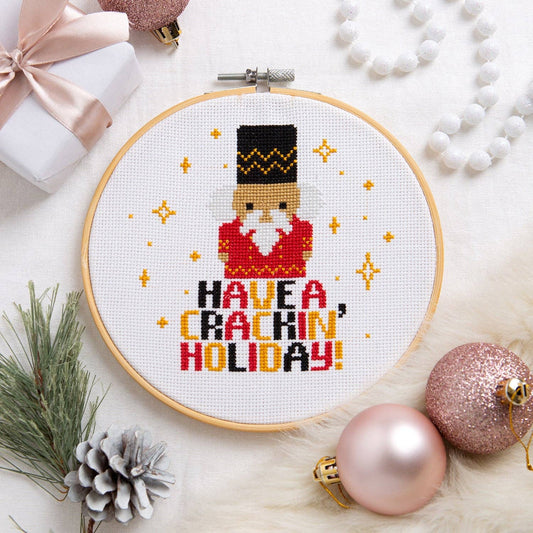 A circular embroidery hoop displays a completed cross-stitch design of a nutcracker with the text 'HAVE A, CRACKIN' HOLIDAY!' surrounded by festive holiday props like ornaments, a pinecone, and a gift box on a white faux fur background.