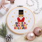 A circular embroidery hoop displays a completed cross-stitch design of a nutcracker with the text 'HAVE A, CRACKIN' HOLIDAY!' surrounded by festive holiday props like ornaments, a pinecone, and a gift box on a white faux fur background.