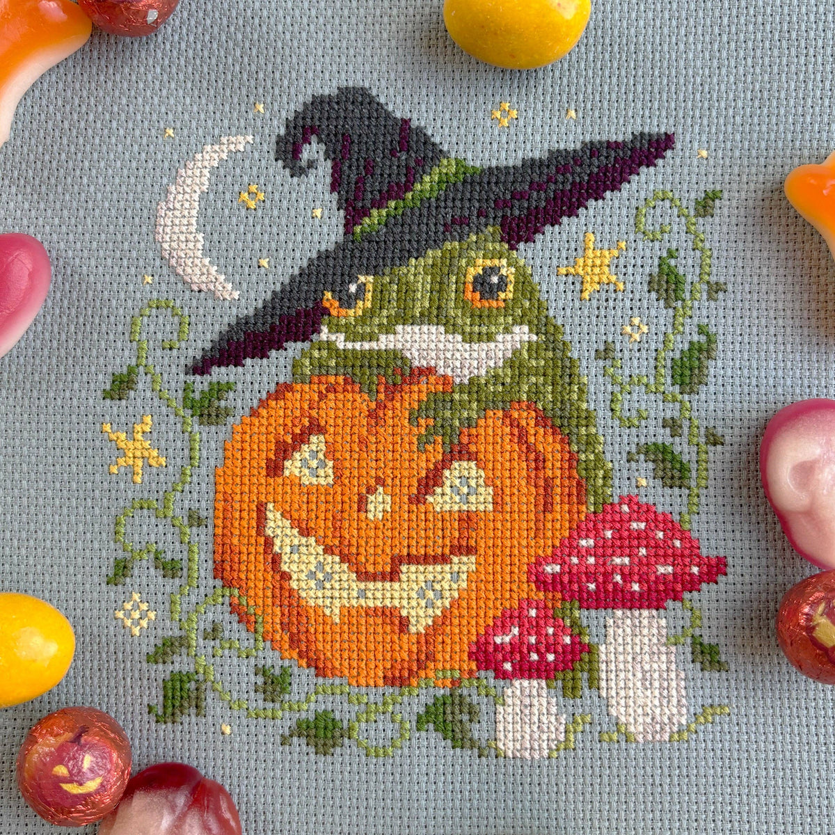 Cross-stitched design of a frog with a witch hat and pumpkin, surrounded by Halloween-themed candies.