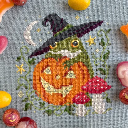 Cross-stitched design of a frog with a witch hat and pumpkin, surrounded by Halloween-themed candies.