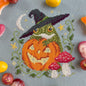 Cross-stitched design of a frog with a witch hat and pumpkin, surrounded by Halloween-themed candies.