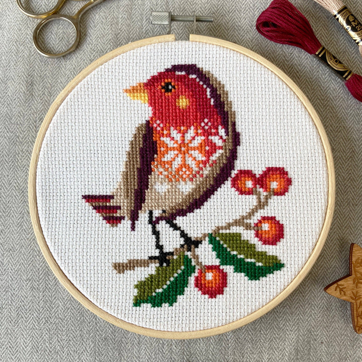Cross-stitched robin on a branch with berries in an embroidery hoop, surrounded by crafting tools.