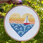 Cross-stitched heart design with a sailboat on fabric inside a wooden hoop, against a green leafy background.