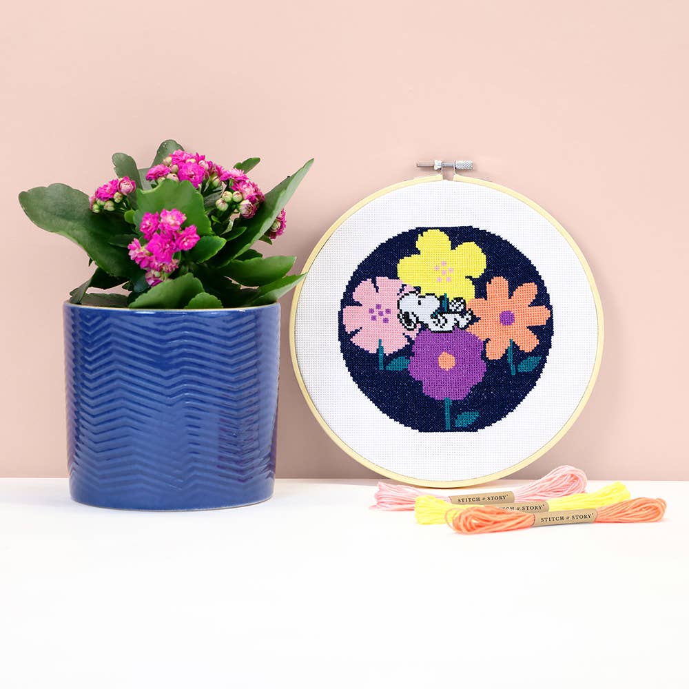 A completed circular cross-stitch embroidery in a wooden hoop showing colorful flowers on a dark navy background, displayed next to a blue potted plant and embroidery floss on a white surface against a pale pink wall.