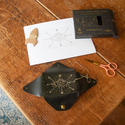 A flat lay of a handmade star sign kit on a wooden table, including a black envelope pouch with gold starry details, a white paper template with a star eye design, a small wooden star, pink-handled scissors, and a black branded box labeled 'Stitch Your Star Sign Envelope Purse'.