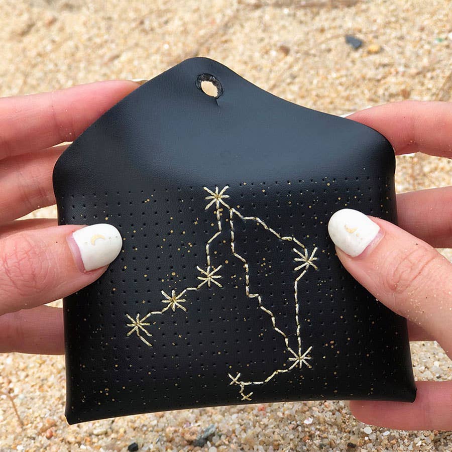 A black leather pouch with a gold constellation design, held by two hands over a sandy beach background.