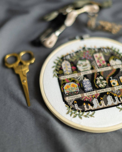 The Apothecary Cross Stitch Kit by Innocent Bones. Cross-stitched scene in an embroidery hoop with gold scissors on a gray fabric background