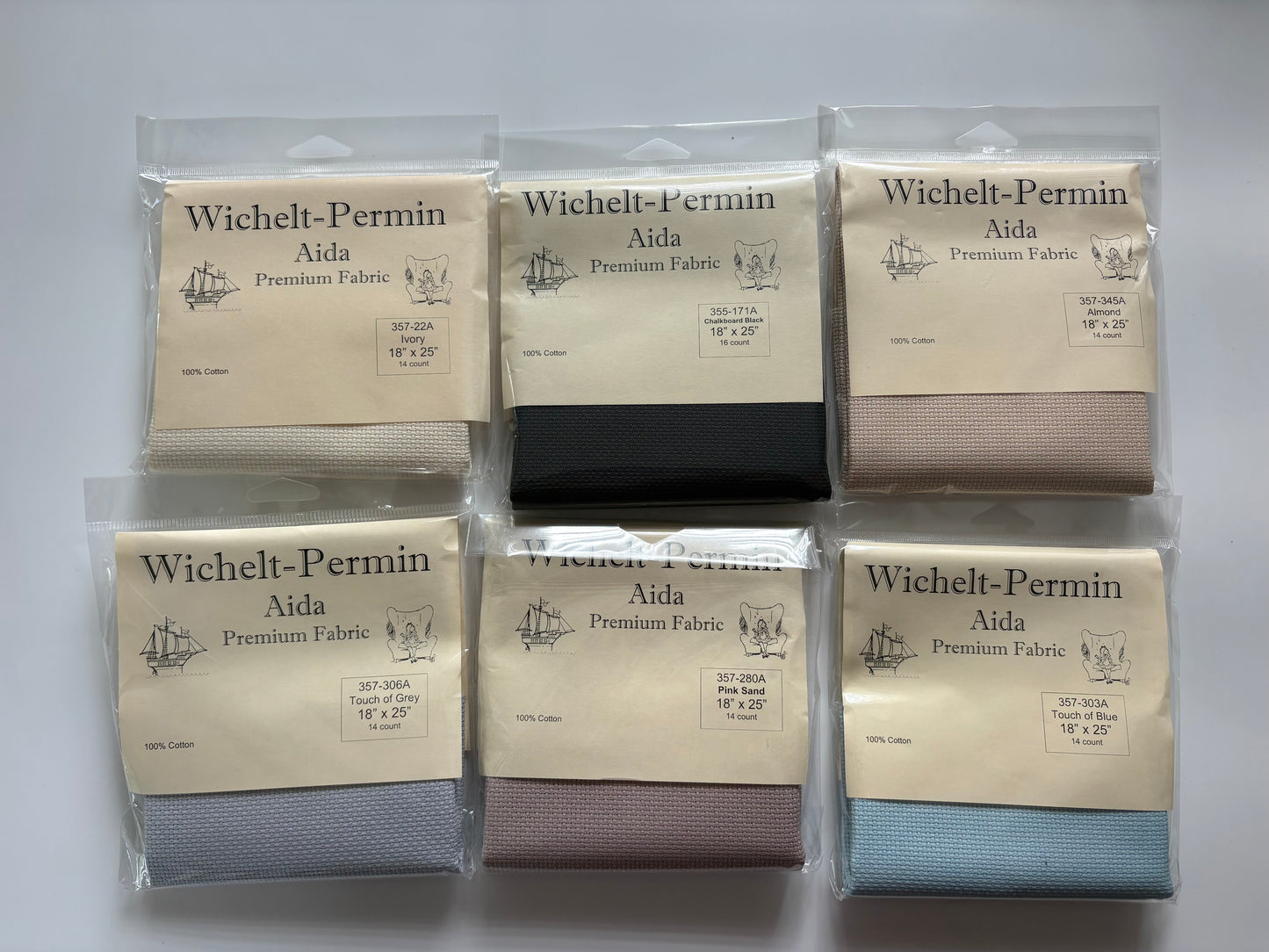 Six packaged fabric bolts from Wichelt-Permin Aida Premium Fabric arranged in a 2x3 grid on a white background. The fabrics shown are in beige, black, taupe, light blue, dusty rose, and navy colors.