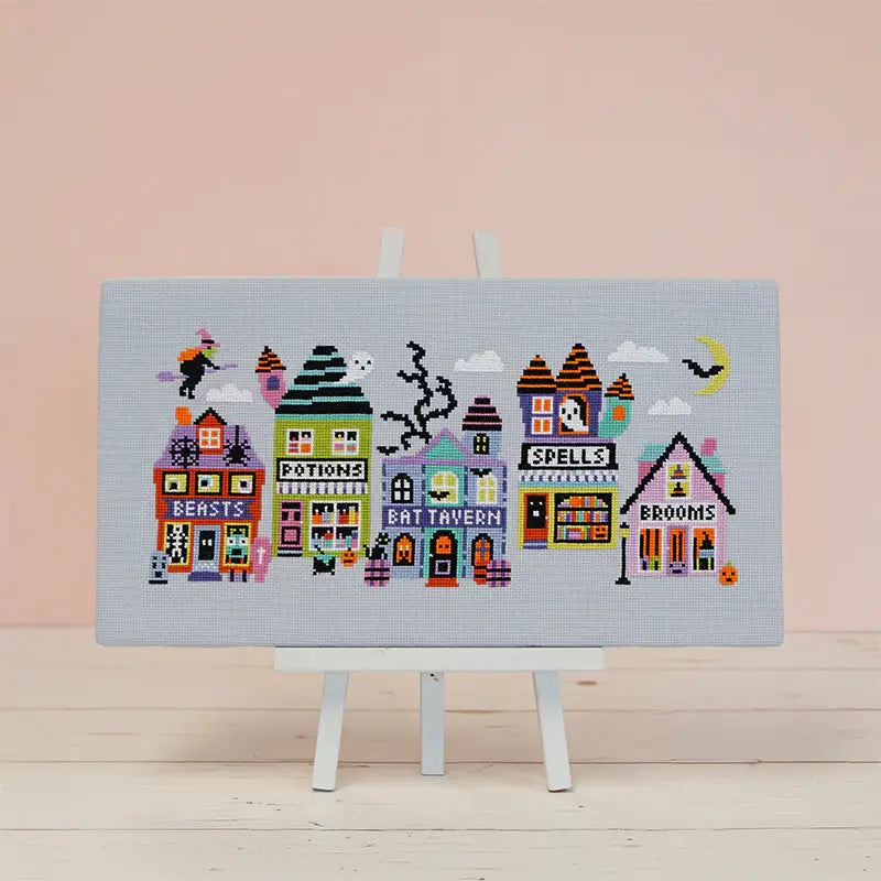 Cross-stitched design of colorful buildings on a light blue fabric with a pink background